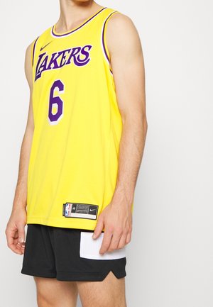 Persona con una camiseta de baloncesto amarilla de los Lakers con el número 6 y pantalones cortos negros con un bolsillo blanco, de pie frente a un fondo liso.