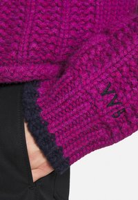 Mano en un bolsillo negro, vistiendo una manga de un suéter de punto grueso color magenta con puño azul oscuro y bordado "WWB".