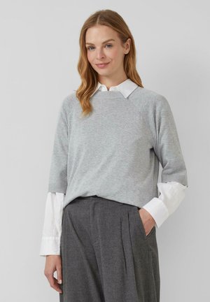 Frau mit hellbraunen Haaren, die einen grauen Pullover über einem weißen Hemd mit Kragen und dunkelgraue, hochgeschnittene Hose trägt, mit einer Hand in der Tasche.