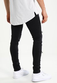 Černé skinny džíny se zipy na stehnech, vyrobené z denimu. Nosené s dlouhou bílou košili a bílými teniskami.