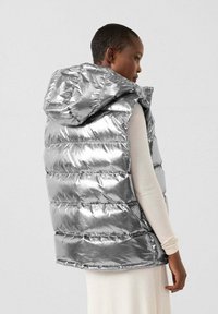 Persona che indossa un piumino argento lucido con cappuccio sopra una maglia beige a maniche lunghe, vista da dietro con un leggero angolo laterale.