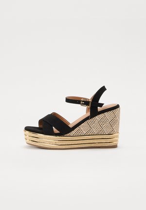 Black suede wedge sandal with a woven natural fiber heel and gold accents. Features cross straps and an adjustable ankle strap.