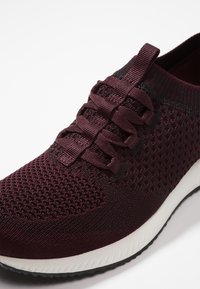 Vinröd sportig sko med en strukturerad stickad ovandel, platta skosnören och en vit mellansula med en svart slitbana.