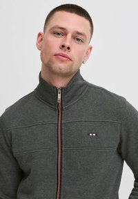 Siva zip-up fleka u uskom dizajnu s narančastim zatvaračem. Ima visoki ovratnik i minimalni logo na prsima. Mekana tekstura.