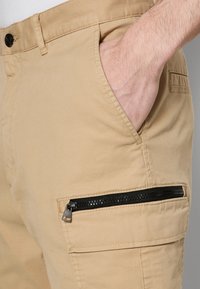 Pantalones cargo color caqui hechos de algodón, con un bolsillo con cremallera y herrajes negros, bolsillos laterales y un acabado liso y texturizado.