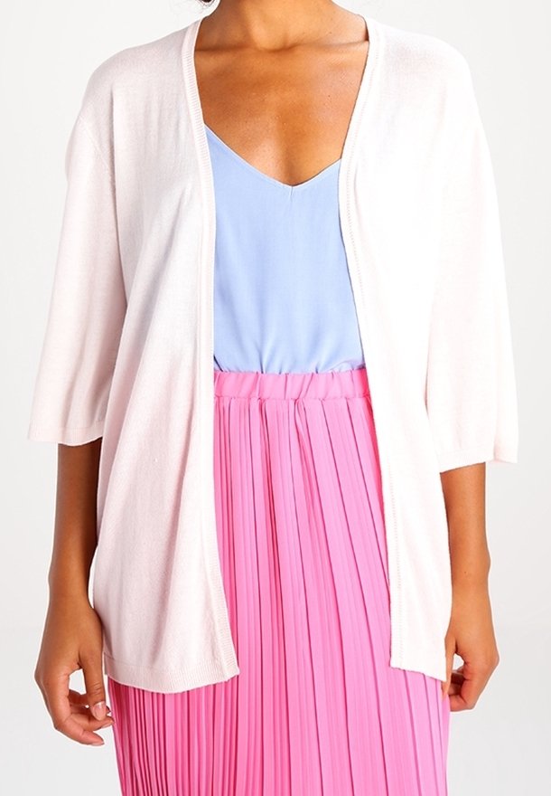 Světle růžový otevřený kardigan s krátkým rukávem, nošený přes modrý top. Modelka ho kombinuje s plisovanou růžovou sukní, která ukazuje jemné materiály.