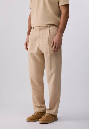Uomo che indossa pantaloni beige, t-shirt beige abbinata e sneakers marrone chiaro, in piedi davanti a uno sfondo chiaro e uniforme.
