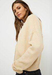 Beige knitted sweater with a relaxed fit, featuring a round neckline and long sleeves, textured fabric, and ribbed hemline.