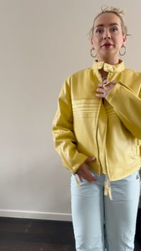 Chaqueta de cuero sintético amarilla con cuello alto, cremallera frontal y detalles de costura horizontal. Combinada con pantalones azul claros y accesorios plateados.