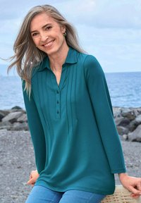 Maglia a maniche lunghe color teal con colletto, bottoni e pieghe verticali. Tessuto morbido con una vestibilità comoda, abbinata a jeans blu chiaro.