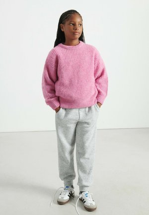 Rosa gestrickter Pullover mit Rippenstruktur, passende graue Jogginghosen und weiße Sneaker mit braunen Sohlen und blauen Akzenten, stehendes Mädchen.