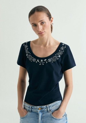 Mujer con una camiseta de manga corta azul marino con bordados blancos florales y de paisley alrededor del escote redondo y vaqueros azul claro.