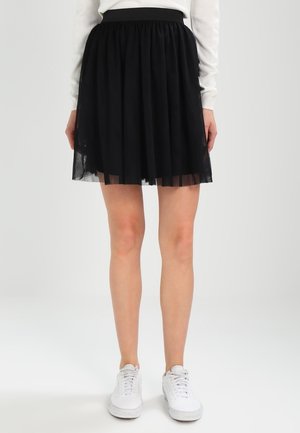 Kvinde iført en sort plisseret nederdel, hvid langærmet top og hvide sneakers, stående mod en ensfarvet lys baggrund.