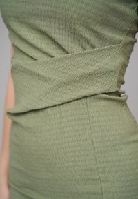 Gros plan sur une robe tricotée verte texturée, ajustée et légèrement froncée autour de la taille sur le torse d’une personne.