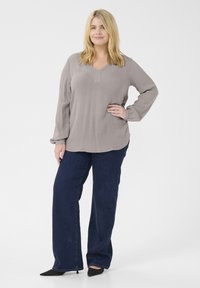 Blouse gris clair à manches longues avec un col en V et des manches légèrement froncées, associée à un jean droit bleu foncé et des chaussures noires à talons pointus.