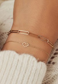 Gouden schakelarmband met een hartbedel en een dunne gouden ketting, met gladde oppervlakken en een gepolijste afwerking, gedragen om een pols met zichtbare huidtextuur.