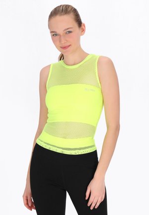 Ung kvinde med blondt hår iført en ærmeløs neongul nettop og sorte bukser, stående mod en hvid baggrund.