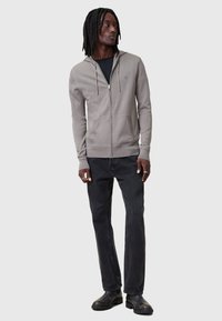 Felpa grigia con zip e texture morbida, caratterizzata da una tasca a marsupio e polsini a coste, indossata sopra una maglietta nera e abbinata a jeans neri.