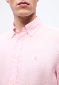 Κοντινό πλάνο ενός ατόμου που φοράει ανοιχτό ροζ πουκάμισο με κουμπιά και γιακά με κουμπί, με ένα μικρό κεντητό λογότυπο στο στήθος.