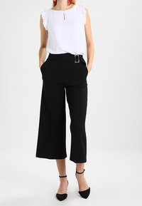 Blusa bianca senza maniche con un motivo texturizzato e maniche a volant, abbinata a pantaloni neri a gamba larga a vita alta e décolleté neri.