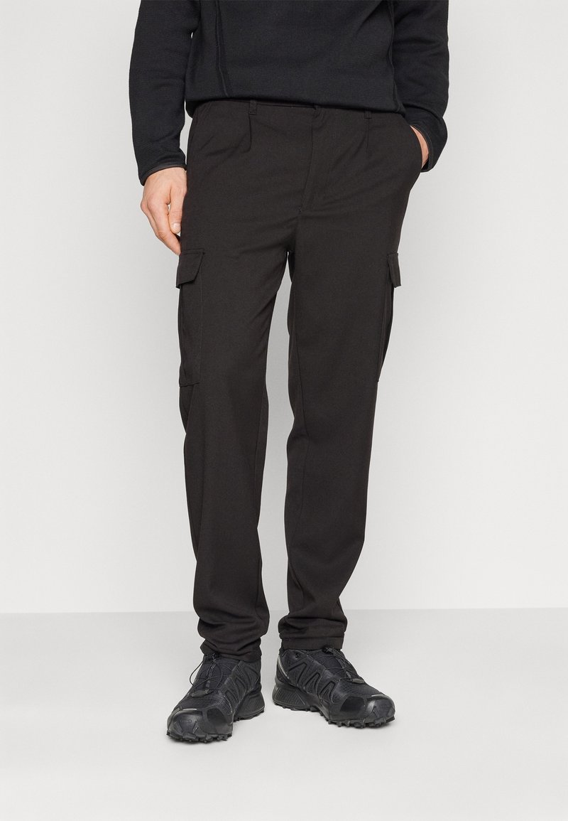 Homme portant un pantalon cargo noir avec des poches latérales et des chaussures de sport noires, se tenant devant un fond clair uni.
