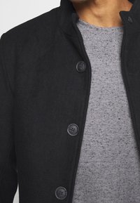 Černý kabát z vlněné směsi s vysokým límcem, s pěti kulatými černými knoflíky a pod ním texturovaný šedý pletený svetr.