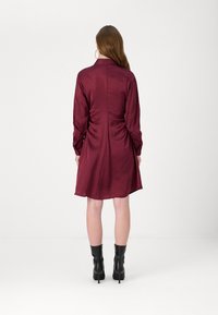 Vestido de seda bordeaux com mangas longas, decote com gola e detalhes franzidos nas costas; combinado com botas pretas até ao tornozelo.