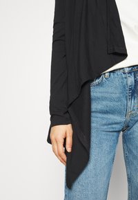 En persons överkropp iklädd en svart långärmad tröja med asymmetrisk nederkant och blå denimjeans mot en vit bakgrund.