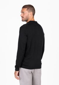 Maglione nero a maniche lunghe con collo rotondo, polsini e orlo a costine, realizzato in un tessuto morbido, esposto di lato su uno sfondo neutro.