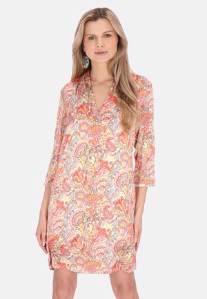 Mujer con un vestido de largo hasta la rodilla, escote en V, con patrones florales de colores rosa, naranja y amarillo en estilo paisley, y mangas tres cuartos, de pie frente a un fondo blanco.