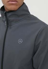 Gray zip-up jacket with high collar, smooth texture, features a small white logo on the left side, showcasing a minimalistic design.