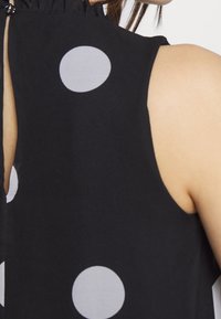 Svart tygtop med stora gråa prickar, med en volangkrage och öppen ryggdesign. Slät textur, avslappnad passform.