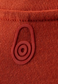 Gros plan sur un tissu tricoté orange rouille avec un emblème en caoutchouc en relief présentant des cercles concentriques à l'intérieur d'une forme ovale.