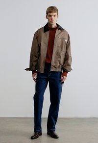 Chaqueta ligera marrón con cuello negro, cremallera en el bolsillo delantero, usada sobre un suéter color óxido y jeans azules, combinada con zapatos marrones.