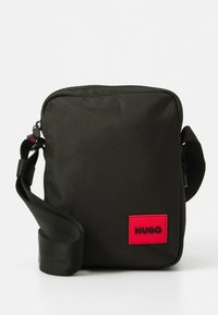 Mala tiracolo preta em nylon com acabamento texturizado, com um remendo de logótipo vermelho contrastante. Inclui uma alça ajustável robusta.