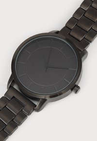 Round black watch with a minimalist design, features a matte dial, thin hour markers, and a black metal link bracelet. Metal buckle is visible.