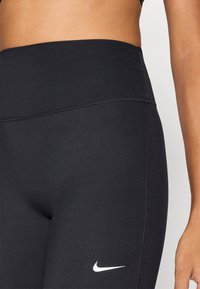 Čierne legíny s vysokým pásom z hladkého, elastického materiálu. Majú ploché švy a malý biely nápis Nike na ľavom stehne.