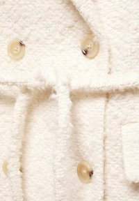 Biely textúrovaný materiál s chlpataným povrchom, s béžovými gombíkmi a viazaním, zvýrazňujúci jemný, teplý dizajn vhodný na vonkajšie oblečenie.