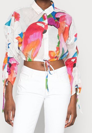 Chemise courte boutonnée avec imprimé coloré de perroquet et floral, associée à un pantalon blanc taille haute.