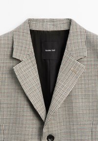 Blazer met een pied-de-poule-patroon in bruin en crème, met een klassieke reverskraag, een enkele zwarte knoop en een zwarte voering.