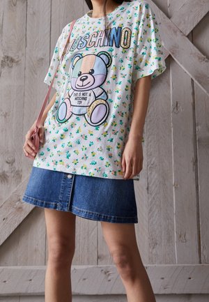 Kobieta nosząca kwiecistą koszulkę z kolorową grafiką misia i tekstem "Moschino", w połączeniu z niebieską dżinsową spódnicą i różową torbą na ramię.
