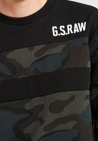 Czarna bluzka z długim rękawem w zielony i niebieski kamuflaż, z prążkowanym okrągłym dekoltem oraz białym napisem "G.S.RAW" na lewym ramieniu. Wykonana z miękkiego bawełnianego materiału.