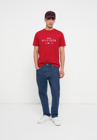 Mladý muž v červeném tričku Tommy Hilfiger, modrých džínách, bílých teniskách a tmavě červené kšiltovce stojící na bílém pozadí.