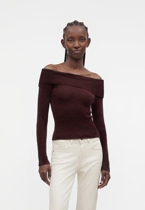 Kvinde med fletninger iført en mørk vinrød sweater med bare skuldre og lyse bukser, der står mod en ensfarvet baggrund.
