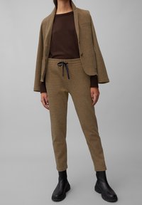 Bruine textuur blazer over een bruin sweater en beige gestreepte joggers met een trekkoord. Zwarte enkellaarsjes maken de outfit compleet.