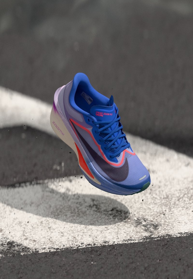 Chaussure de course Nike Zoom Fly bleue et violette avec des accents orange flottant au-dessus d’un bitume avec des marquages routiers blancs.
