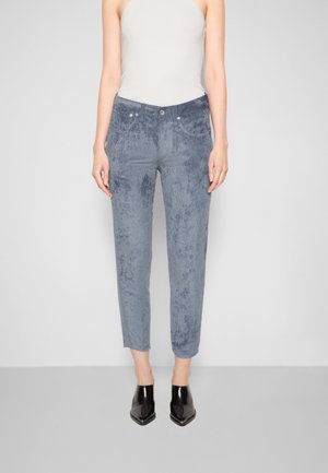 Frau trägt graue Cordhose mit hohem Bund, schwarze Stiefeletten mit spitz zulaufender Spitze und ein ärmelloses weißes Oberteil, steht vor einem schlichten Hintergrund.