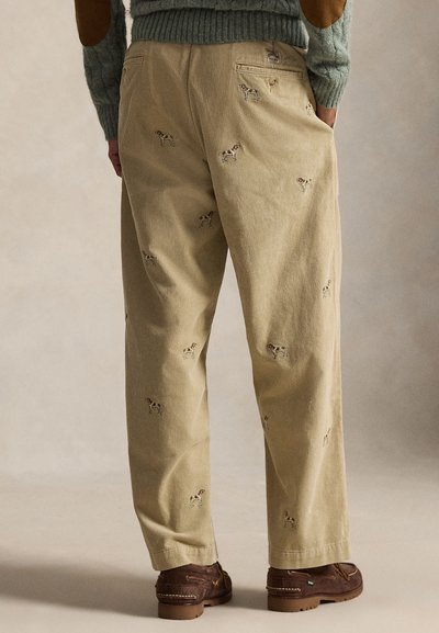 Pantaloni in velluto a coste beige con ricamo di cane, taglio dritto e tasche posteriori. Abbinati a scarpe marroni senza lacci e un maglione verde a maglia.