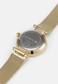 Montre avec bracelet en maille dorée, boîtier rond, dos en acier inoxydable gravé "LACOSTE" et étanchéité jusqu'à 3 bars.