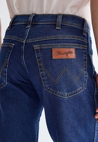 Gros plan d'une personne portant un jean Wrangler bleu foncé montrant la poche arrière avec un patch en cuir marron et des coutures jaunes.